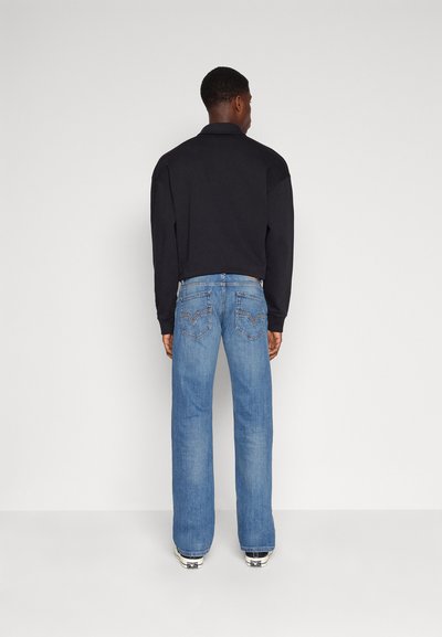 Un homme à la peau foncée portant une chemise noire à manches longues, un jean droit bleu et des baskets blanches se tenant devant un mur blanc.