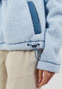 Lichtblauwe fleecetrui met gestructureerde stof, blauwe ritszak, verstelbare trekkoord onderkant, gedragen over beige corduroy broek en een hand ontspannen aan de zijkant.