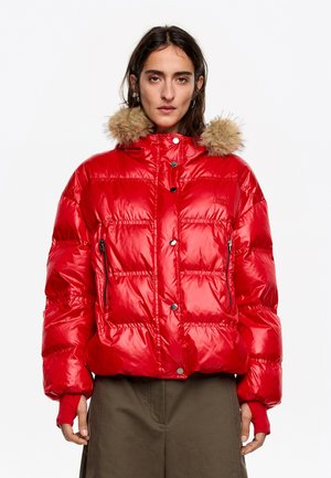 Czerwona kurtka puchowa o połyskującym wykończeniu, z kapturem wyłożonym futrem, kieszeniami na zamek i zatrzaskami. Krótki, pikowany fason.