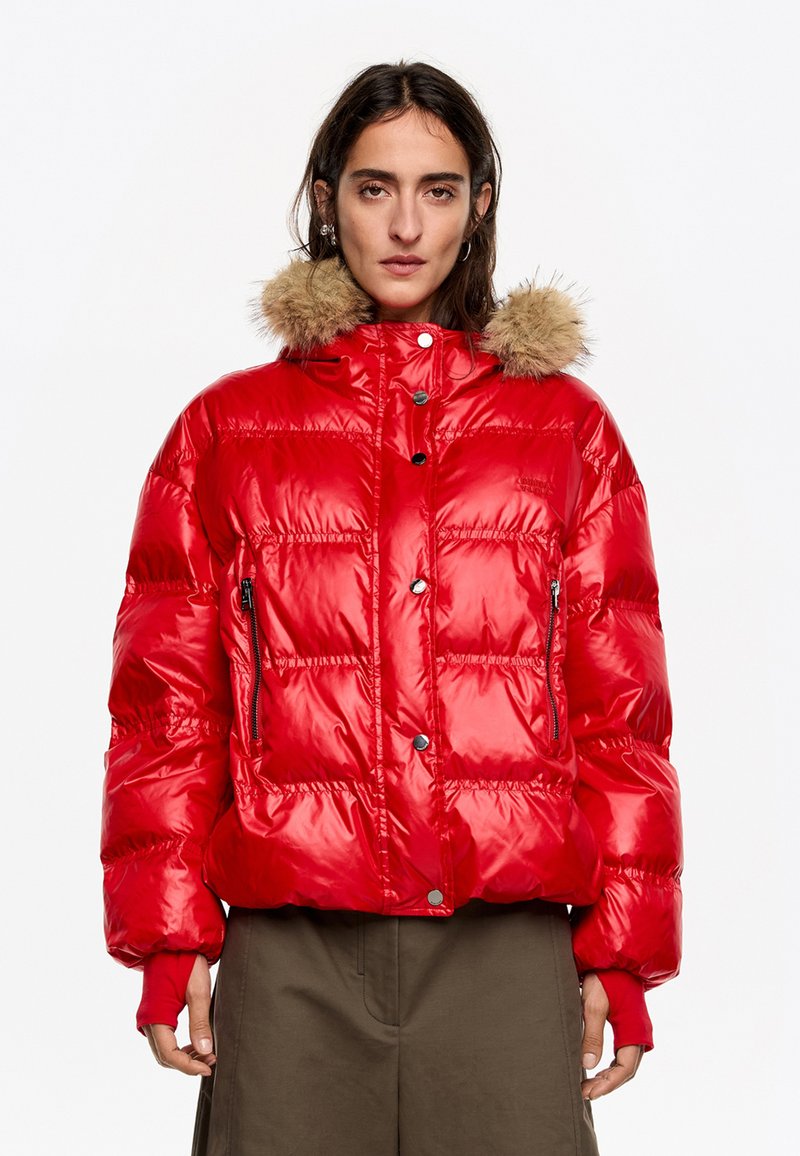 Czerwona kurtka puchowa o połyskującym wykończeniu, z kapturem wyłożonym futrem, kieszeniami na zamek i zatrzaskami. Krótki, pikowany fason.