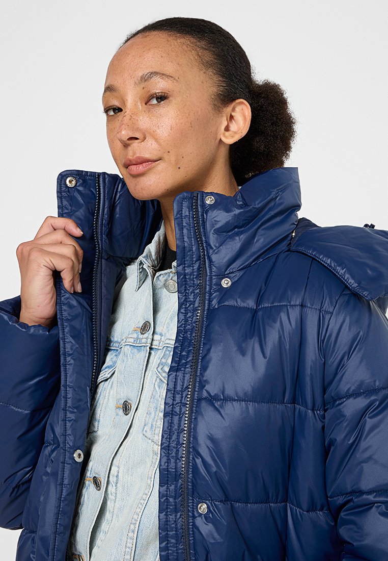 Giacca puffer blu navy con colletto alto, bottoni a scatto e texture trapuntata. Indossata sopra una camicia in denim chiaro, che mostra uno stile stratificato.
