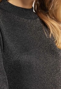 Gros plan sur une personne portant un haut en tricot noir pailleté avec un col côtelé, montrant une partie de son cou, de son épaule et une grande boucle d'oreille.