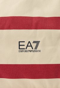 Platnena torba s krem pozadinom i smjelim crvenim prugama. Na njoj se ističe "EA7 EMPORIO ARMANI" logo u crnoj boji. Tekstura tkanine.
