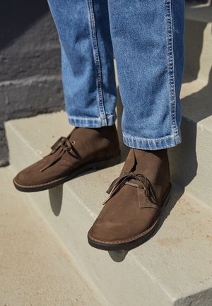 Bottes désert en daim marron à lacets portées avec un jean en denim bleu, debout sur des marches en béton gris clair sous la lumière du soleil.