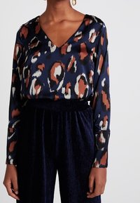 Marinblus med v-ringning och långa ärmar, prydd med ett leopardsmönster i bruna, vita och rostfärgade nyanser. Texturerad tyg och avslappnad passform.