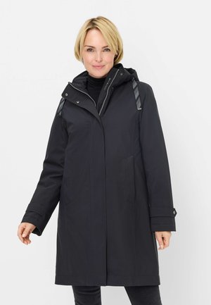 Frau trägt einen schwarzen, langen Kapuzenmantel über einem schwarzen Rollkragenpullover, steht vor einem schlichten weißen Hintergrund und blickt nach vorn.