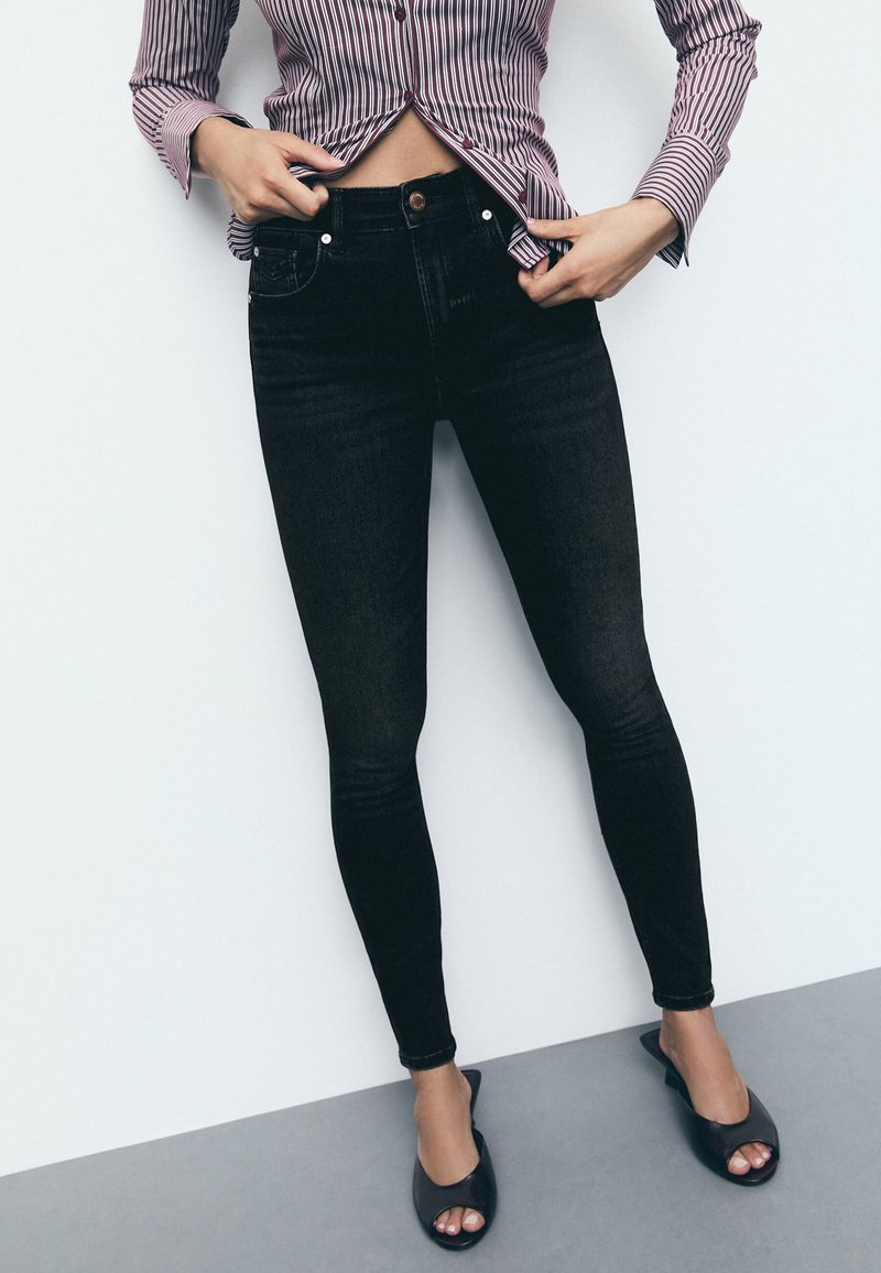 Personne portant un jean skinny noir, des talons ouverts noirs et une chemise à manches longues rayée, relevant l'ourlet pour montrer la taille.