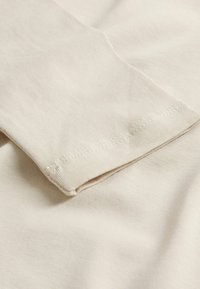 Beige bomullstyg med slät yta, med en vikt ärm med sydd fåll. Mjukt, lätt material visas i närbild.
