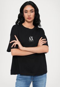 Juodas medvilnės marškinėliai su trumpomis rankovėmis, su baltu logotipu "AX" priekyje ir laisvu pritaikymu. Dėvimi su mėlynomis džinsomis.