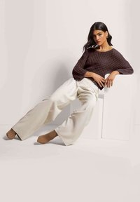 Brauner gestrickter Pullover mit strukturiertem Muster, kombiniert mit weiten beigen Hosen. Das Modell posiert vor einem neutralen Hintergrund und sitzt auf einem weißen Block.