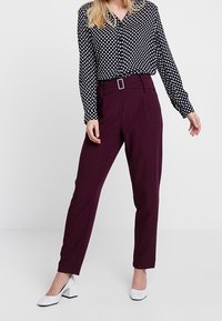 Blusa bianca e nera a pois con bottoni, abbinata a pantaloni bordeaux a vita alta con passanti per cintura frontali. Scarpe con tacco a blocco bianche.