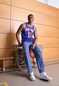Blå basketballtrøje med orange detaljer og "NEW YORK" tryk, parret med lyse denimjeans og hvide sneakers. Kurve i baggrunden.