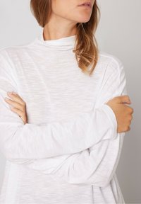 Bijela majica dugih rukava s lažnim visokom ovratnikom, izrađena od lagane, teksturirane tkanine i slobodnim rukavima, s prekriženim rukama na neutralnoj pozadini.
