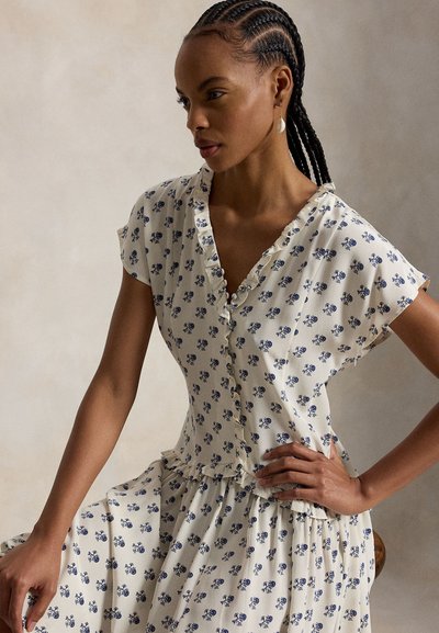 Letitukkaiseen naiseen, joka on pukeutunut valkoiseen mekkoon, jossa on pieniä sinisiä kukkakuvioita ja röyhelöreunukset, istuu käsi lantiolla.