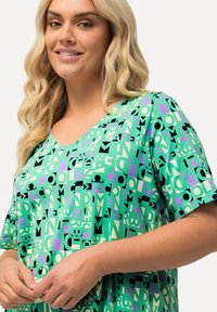 Vihreä lyhythihainen tee, jossa on mustien, purppuraväristen ja kermaisten geometristen kirjainten sekä muotojen sekoitus. Pehmeä kangas, rento istuvuus ja V-pääntie.