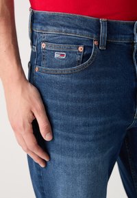 Temno modri kavbojke iz denima z sprednjimi žepi, bakrenimi dodatki in majhnim blagovno znamko oznako z rdečimi, belimi in modrimi elementi na žepu.