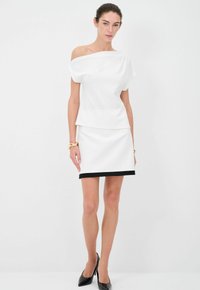 Top bianco off-the-shoulder con design a strati, abbinato a una gonna aderente coordinata con orlo nero, accompagnato da tacchi a punta neri.