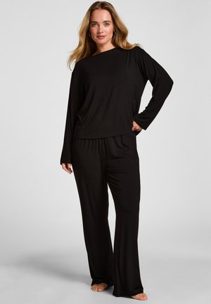 Vrouw met lang blond haar, een losse zwarte top met lange mouwen en een wijde broek, staat op blote voeten tegen een effen grijze achtergrond.