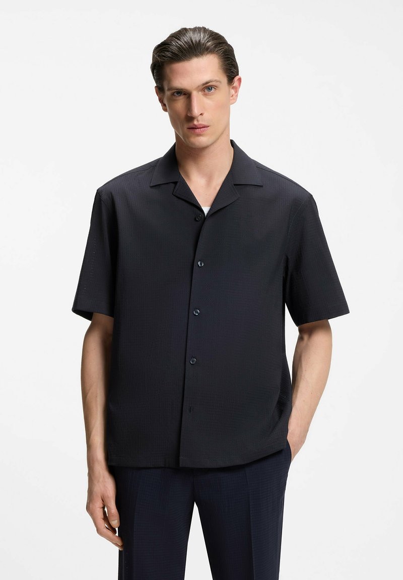 Homme aux cheveux coiffés en arrière portant une chemise noire à manches courtes boutonnée et un pantalon noir assorti, debout avec une main dans la poche.