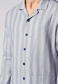 Camicia a righe pulsante in azzurro chiaro e bianco, con colletto, bottoni blu e due tasche frontali in tessuto testurizzato.