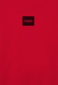 Tissu rouge avec un patch rectangulaire noir centré près de l'encolure affichant le mot « HUGO » en lettres rouges.