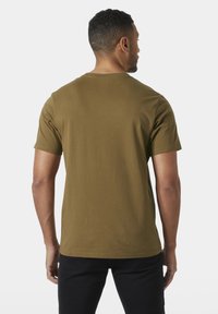 Hombre con cabello corto y rizado, vistiendo una camiseta de manga corta color verde oliva y pantalones negros, de pie mirando hacia atrás contra un fondo claro.