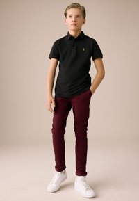 Polo noir à manches courtes, pantalon bordeaux slim-fit, et baskets blanches. Petit logo sur la poitrine du polo.