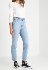 Jasnoszara koszula z długim rękawem, jasnoniebieskie, wysokie dżinsy z detalami typu distressed oraz czarne trampki z białymi akcentami.
