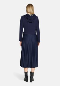 Granatowa bluzka z kapturem o krótkim kroju w zestawieniu z plisowaną granatową spódnicą. Strój ma miękką fakturę i sięga do połowy łydki. Całość dopełniają czarne botki za kostkę.