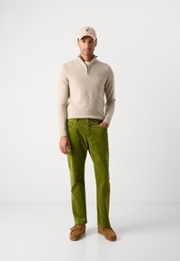 Beige zip-up sweater, grønne corduroy bukser og brune loafers. Kasketten er beige med et lille logo. Modellen står foran en ensfarvet baggrund.