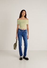 Svetlo zelena ribasta kratka majica z odkritimi rameni, kombinirana z modrimi jeans hlačami. Model drži majhno olivno zeleno torbico, obuje črne čevlje z belimi poudarki.