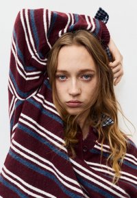 Pulover tricotat cu dungi bordo, cu linii albe și albastre, cu guler și detaliu cu nasturi, purtat peste o cămașă în carouri albastre.