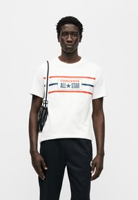 Άσπρο βαμβακερό t-shirt με κόκκινες και μπλε οριζόντιες ρίγες και το λογότυπο «CONVERSE ALL STAR». Μαύρη τσάντα ώμου. Μαύρο παντελόνι.