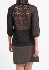Zwarte doorschijnende blouse met driekwartmouwen, kant achterkant met golfpatronen, gecombineerd met een donkergroene rok. Geweven stoffen details zichtbaar.