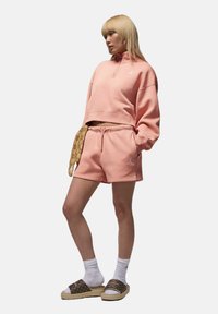 Ljusrosa, kort sweatshirt med halvmundering, tillsammans med matchande shorts. Har Jordan-logotypen. Bärs med vita strumpor och bruna slip-on sandaler.