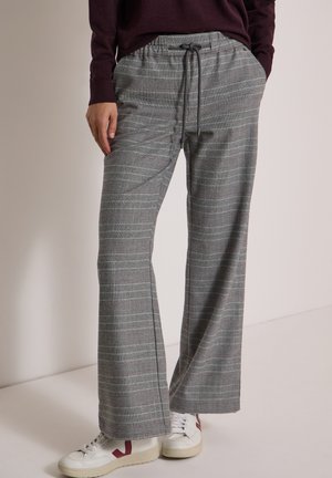 Personne porte un pantalon large à carreaux gris avec un cordon noir et des baskets blanches avec des touches bordeaux, debout devant un fond uni.