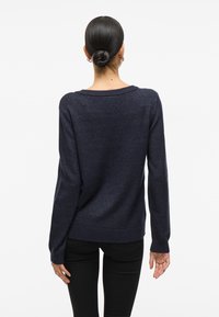 Pull en maille bleu marine avec un large col, des manches longues et un ourlet côtelé. Texture lisse et coupe légèrement ample, porté avec un pantalon noir.