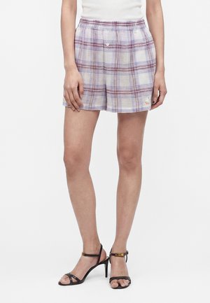 Žena nosí svetlofialové kárované šortky so zapínaním na bočnom gombíku a čierne sandále na podpätku s remienkami okolo členku.