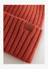 Gorro de punto naranja acanalado con un acabado texturizado y una etiqueta de tela marrón con un diseño de montaña, que destaca por su calidez y detalle de diseño.