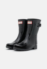 Bottes de pluie en caoutchouc noir avec une finition brillante, bout rond et détail de boucle. Présente une étiquette de marque près du bord supérieur. Semelle texturée.
