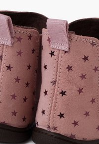 Różowe zamszowe botki za kostkę z wzorem w gwiazdy, z ciemniejszymi różowymi gwiazdami i materiałowymi pętlami ułatwiającymi zakładanie. Brązowe gumowe podeszwy zapewniają przyczepność.