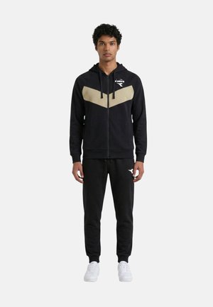 Giovane uomo in piedi che indossa una felpa nera con zip e motivo beige a chevron e pantaloni da jogging neri con scarpe da ginnastica bianche su sfondo semplice.