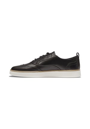 Sneaker in pelle nera con punta a wingtip, suola in gomma bianca e dettagli brogue, visto di lato su uno sfondo bianco.