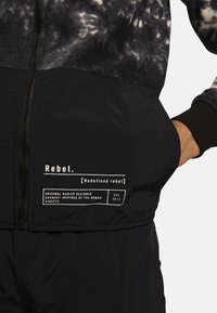 Giacca nera con zip, caratterizzata da una parte superiore tie-dye, tessuto testurizzato e un'etichetta bianca con dettagli di brand e design sulla parte anteriore.