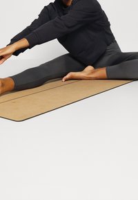 Persona con sudadera negra y leggings estirando una pierna hacia adelante sobre una esterilla de yoga marrón contra un fondo blanco.