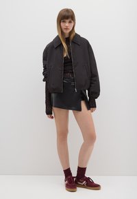 Chaqueta bomber negra con cremallera frontal, blusa de encaje de manga larga, falda envolvente de cuero negro y zapatillas burdeos con acentos blancos y suelas marrones.