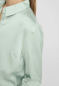 Light green blouse with a soft satin finish, featuring a collared neck, button closure, and puffed sleeves. Smooth texture with subtle sheen.