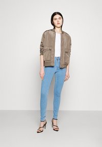 Casaco bomber de cetim castanho com punhos em canelado e dois bolsos frontais, usado sobre uma blusa branca, combinado com jeans skinny azul claro e saltos pretos.