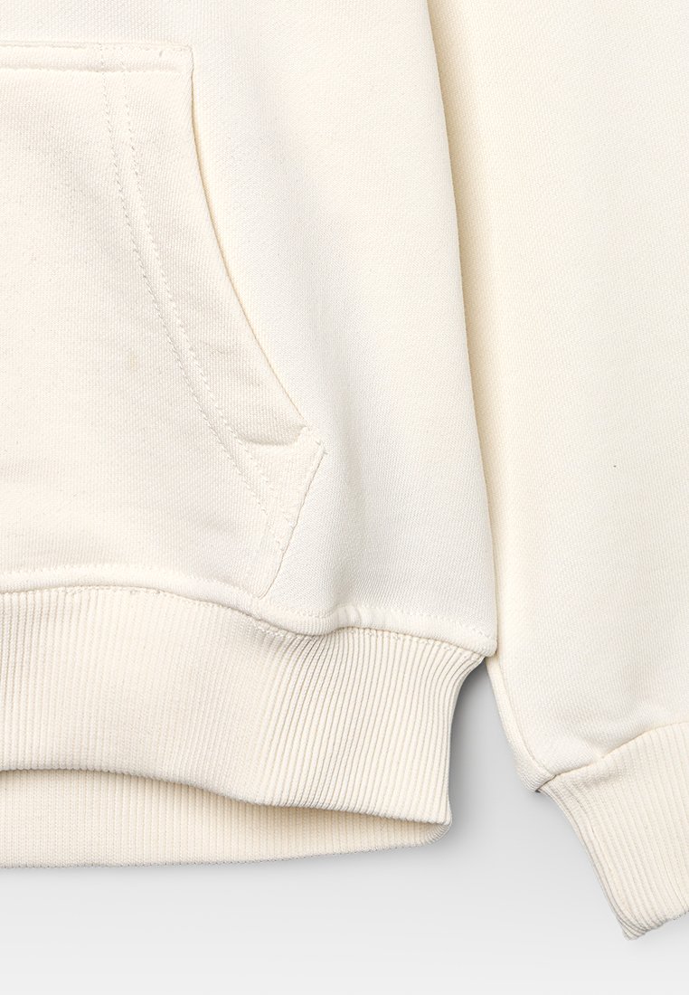 Primo piano di una felpa color crema che mostra i dettagli di cucitura del polsino a costine, della vita e della tasca centrale a forma di canguro.