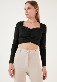 Top corto nero a maniche lunghe con scollatura quadrata e dettaglio frontale arricciato, abbinato a pantaloni beige a vita alta con un bottone dorato.